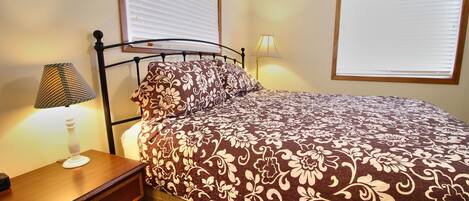 Una cama bien tendida con una colcha estampada con flores, una lámpara de noche y una mesita de noche con un cajón.