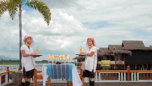 Two individuals serving drinks at a table with a view of a water body and thatched-roof structures.