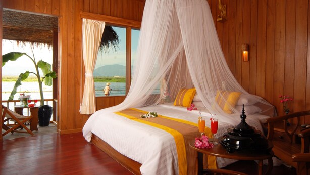 A wooden room with a bed, a table with a drink, and a view of a body of water.