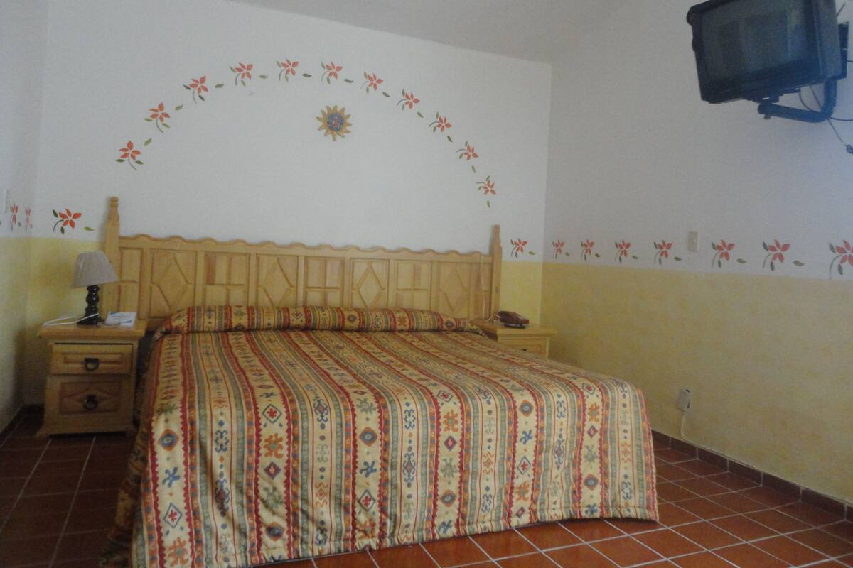 Ein Schlafzimmer mit einem Bett, einem Nachttisch, einer Lampe, einem an der Wand montierten Fernseher und floralen Wandbildern.