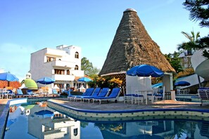 Ein Schwimmbecken mit Liegestühlen und Sonnenschirmen, einer kegelförmigen Struktur und einem Gebäude mit weißer Fassade.