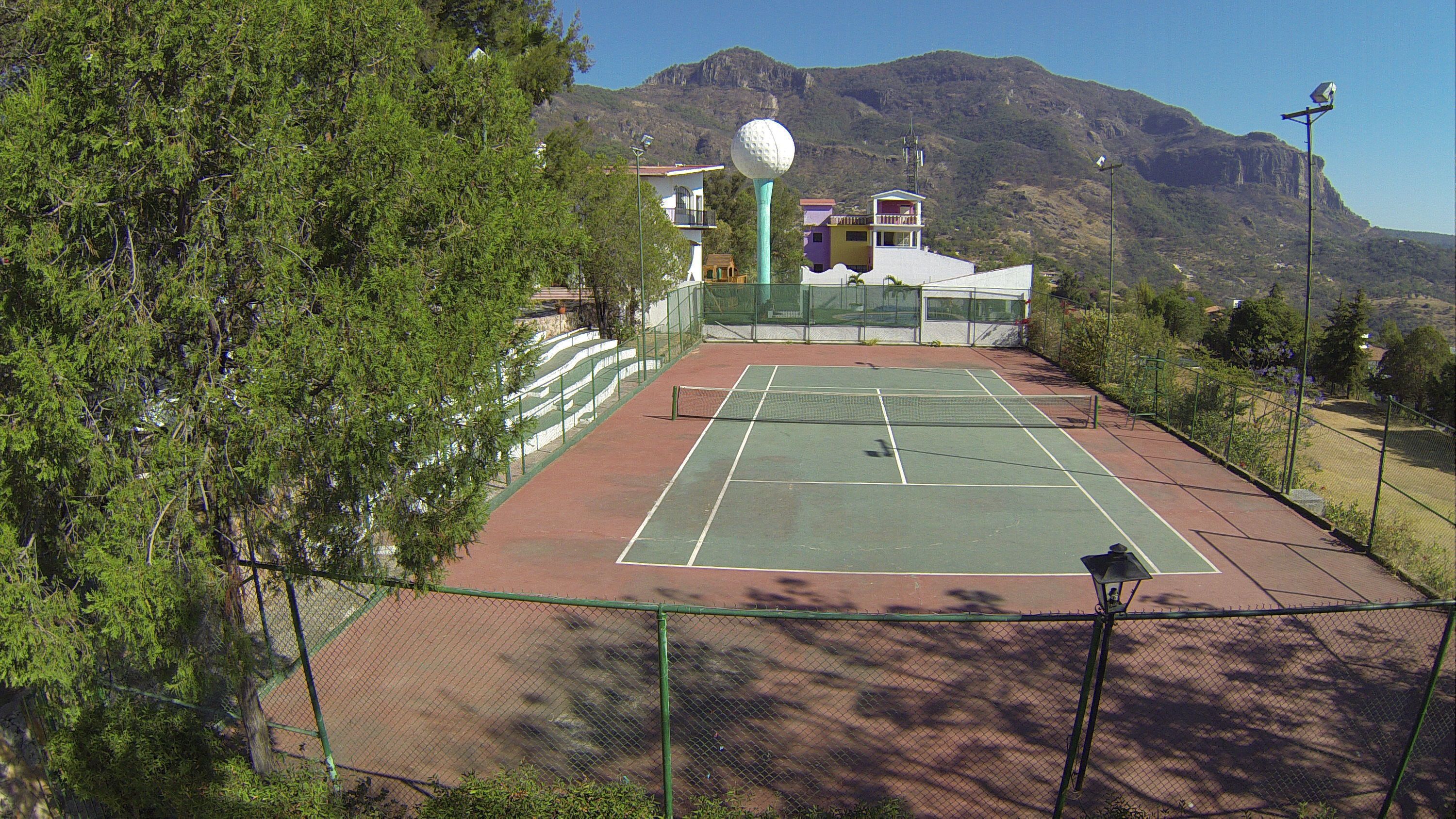 Una cancha de tenis con una red, rodeada por una valla, y un objeto esférico grande al fondo.