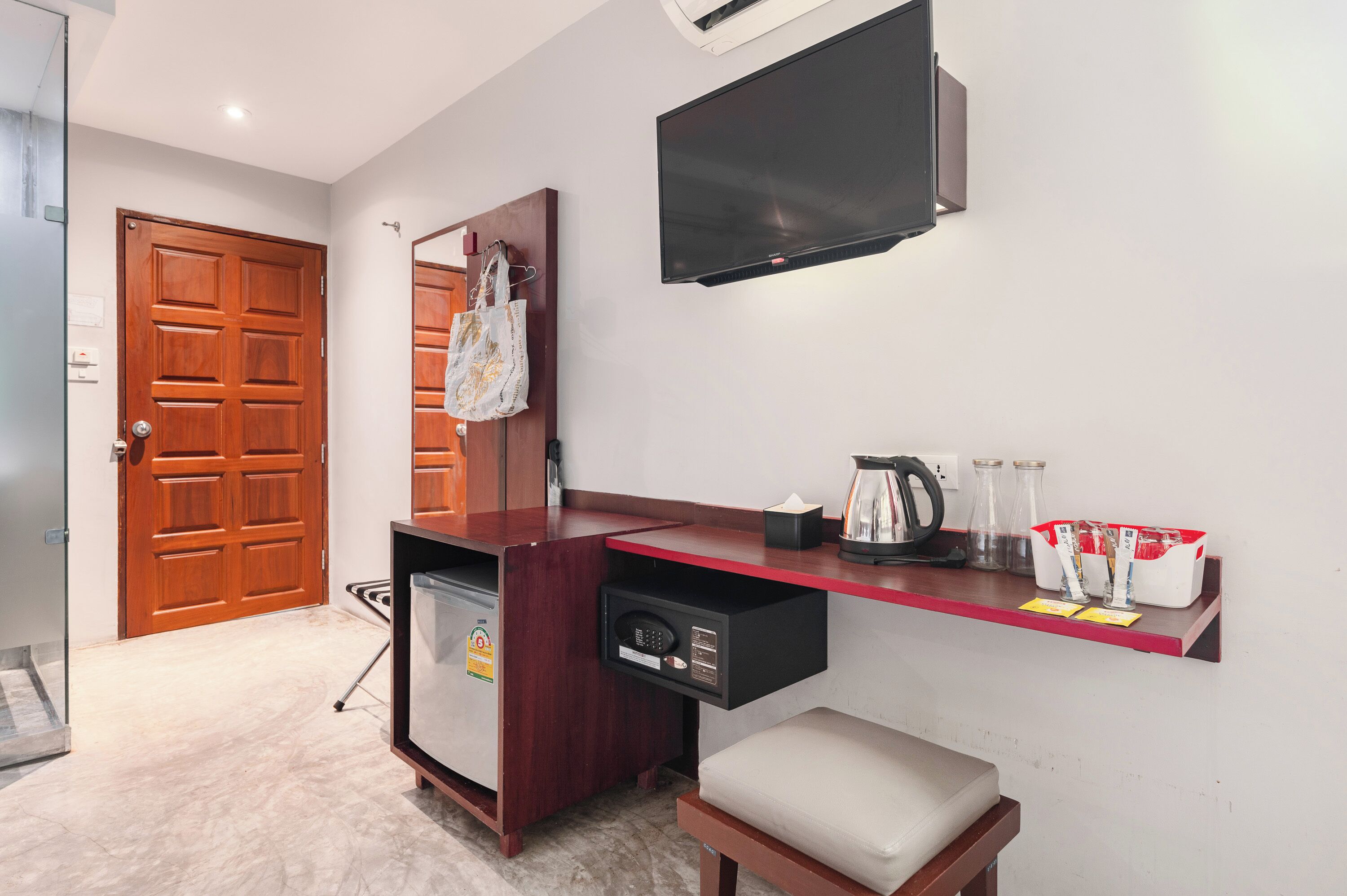 A hotel room with a wooden door, a desk with a computer, a TV mounted on the wall, and a small seating area.