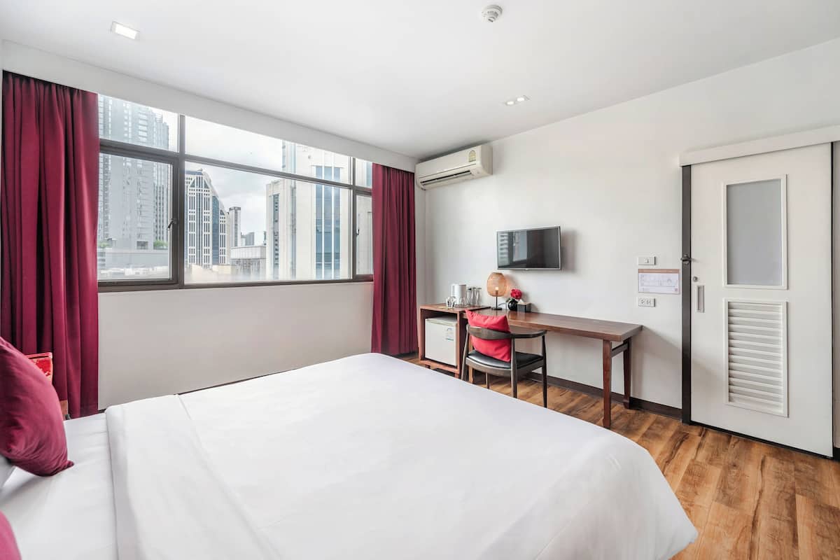 A hotel room with a large bed, a desk with a chair, a television, and an air conditioning unit.