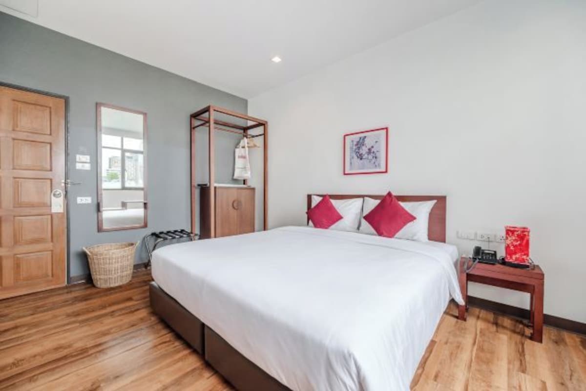 Une chambre avec un plancher en bois, un lit avec une literie blanche et des oreillers rouges, une table de chevet en bois, une armoire en bois et un tableau encadré accroché au mur.