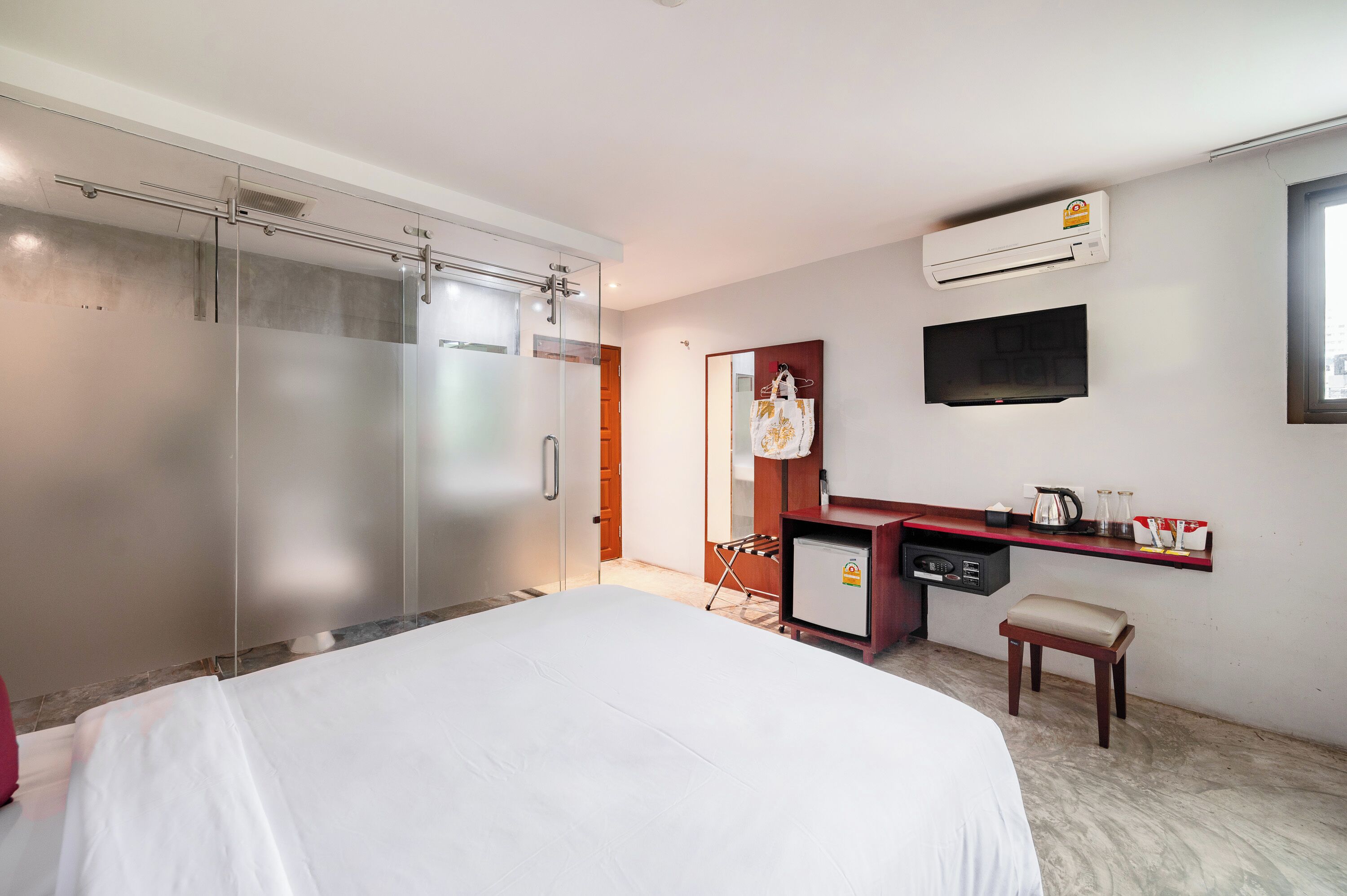 A hotel room with a bed, a desk with a chair, a small refrigerator, a microwave, an air conditioning unit, and a television on the wall.