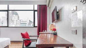 A hotel room with a bed, a wooden table, a chair, a window with a city view, a red curtain, and a wall-mounted TV.