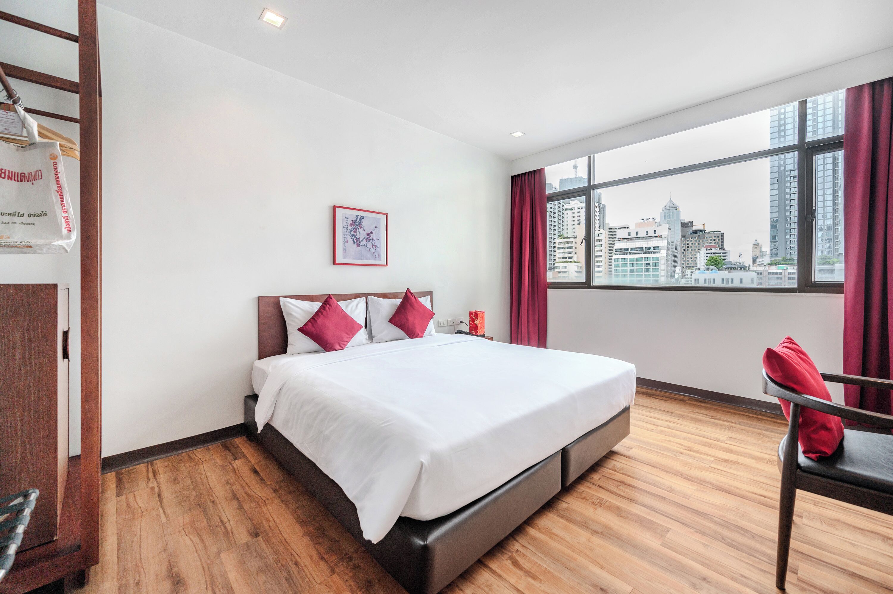 Een hotelkamer met een groot bed, een houten vloer, uitzicht op de stad en een rode stoel.