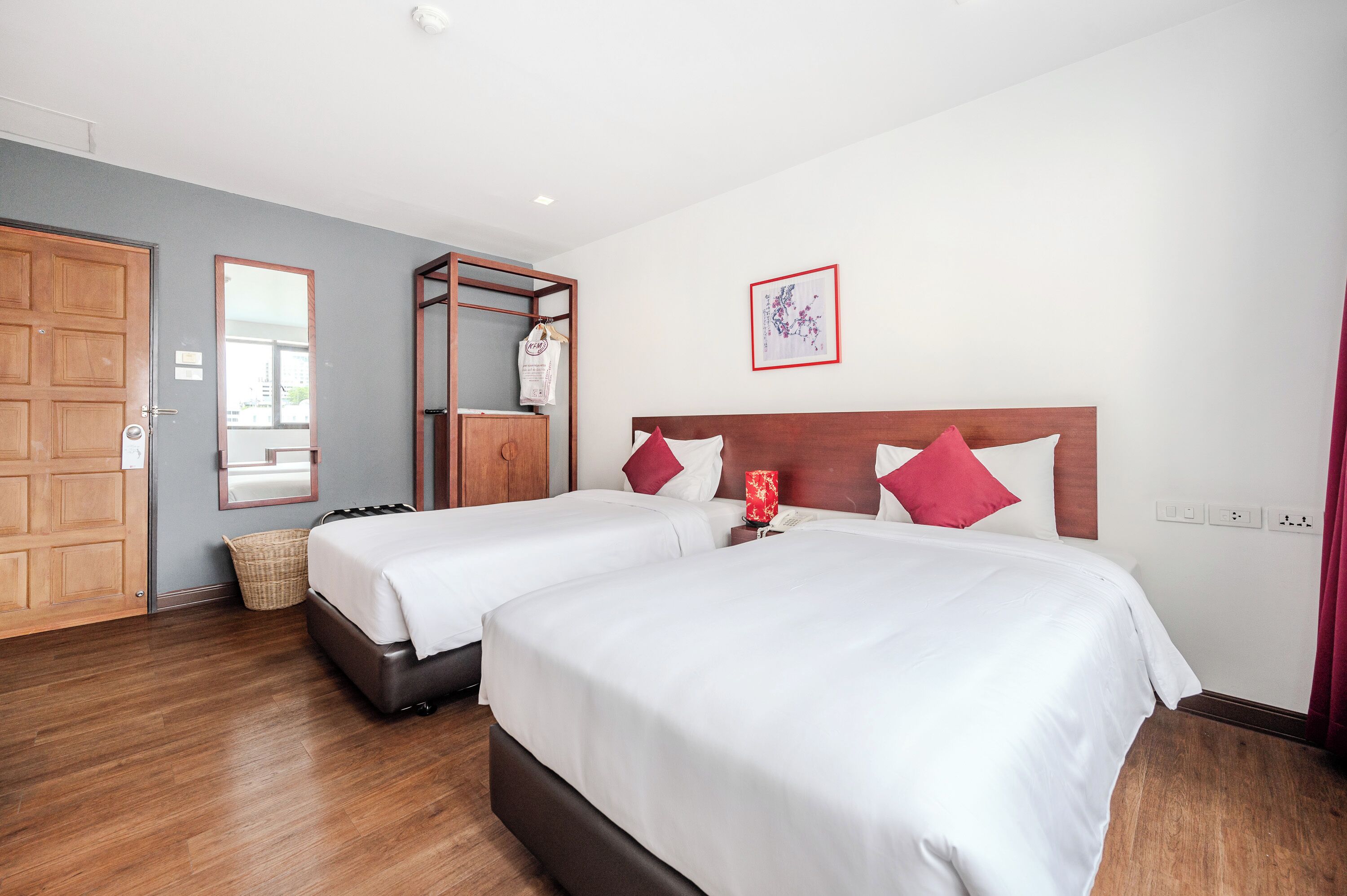 Habitación de hotel con dos camas, cabeceros de madera, un armario de madera, un espejo, un cuadro enmarcado y una lámpara roja.