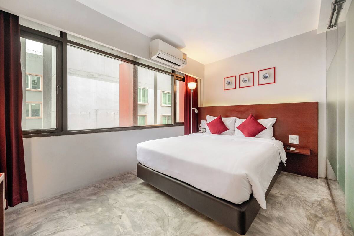 A hotel room with a bed, a window, an air conditioning unit, and a wall-mounted light fixture.