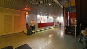 A hotel reception area with a red counter, closed shutters, and a staircase.