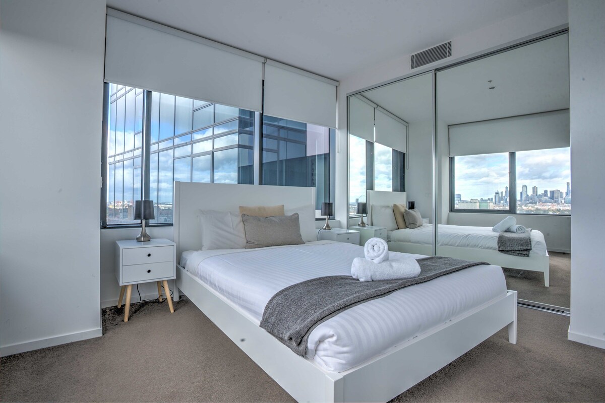 A modern hotel room with a large bed, a nightstand, a mirror, and a view of the city skyline through large windows.