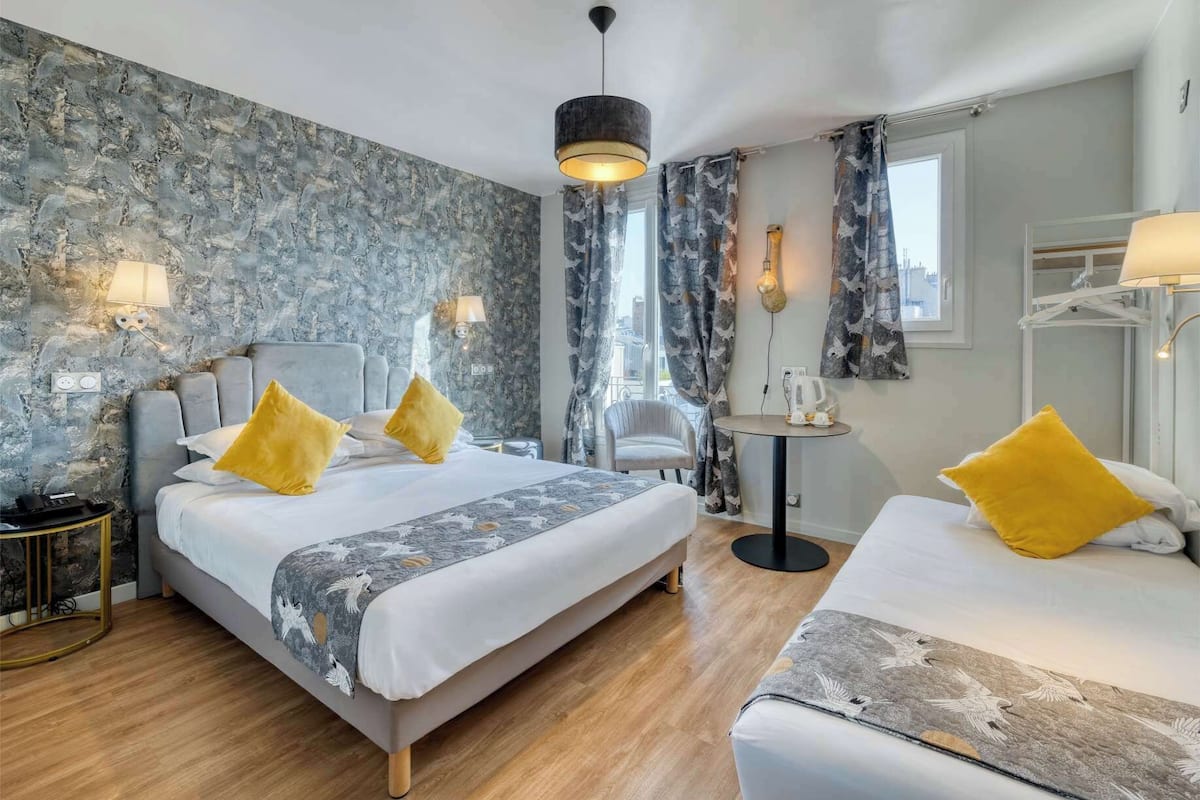 A hotel room with a large bed, a smaller bed, two bedside tables, a chair, a round table, and a view of the city through the window.