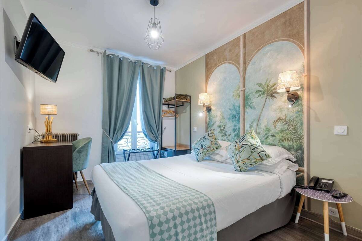 A hotel room with a bed, a desk with a lamp, a TV mounted on the wall, and a window with curtains.