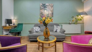 A living room with a grey sofa, a round coffee table, and purple and pink chairs. There is a painting on the wall and a lamp on the right. - Hotel de Belfort (Paris)