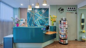 A reception area with a teal counter, a clock, a bulletin board, and a mirror on the wall. - Hotel de Belfort (Paris)