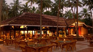 An outdoor restaurant with wicker furniture and a thatched roof, surrounded by palm trees. - Club Bentota (Bentota)