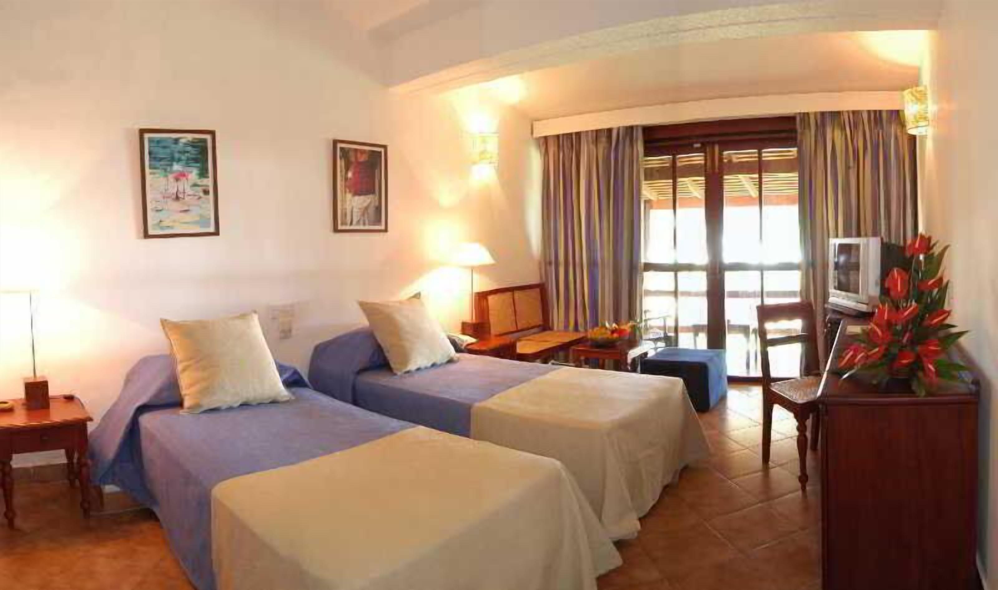 A hotel room with two beds, a TV, a desk, and a window with curtains.