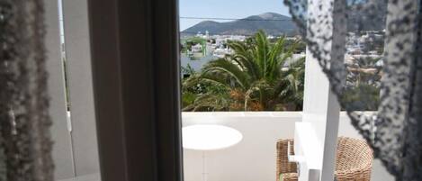 A balcony with a white table and wicker chairs, overlooking a coastal area with buildings and a mountain.