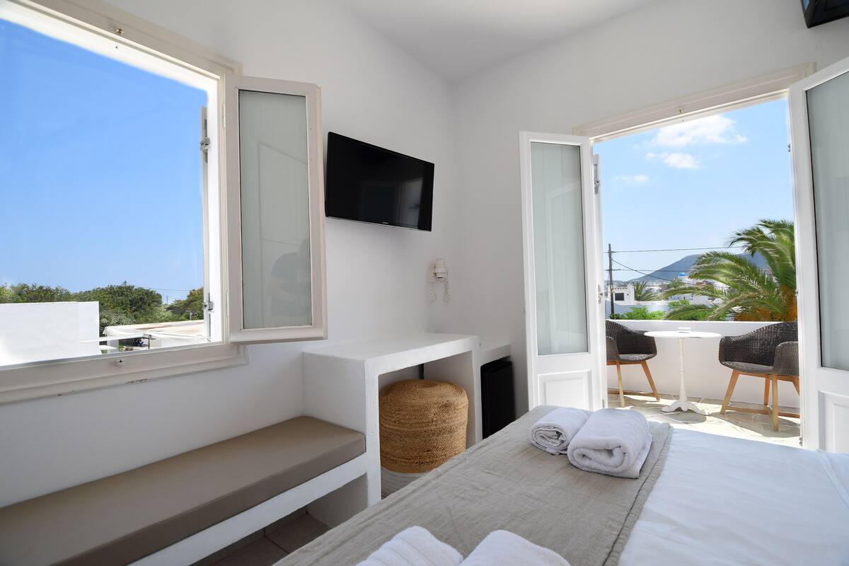 A hotel room with a bed, a TV mounted on the wall, a small table with chairs, and a balcony with a view of palm trees and buildings.