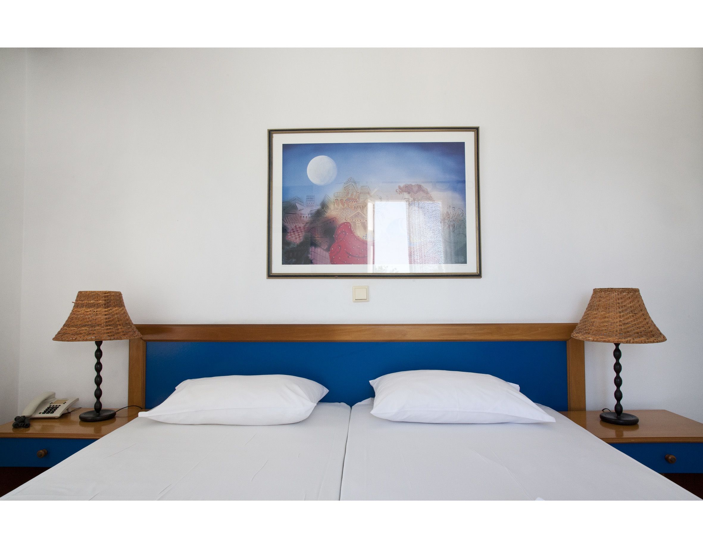 A double bed with white linens, a wooden headboard, two bedside lamps, a framed picture of a landscape with a moon, and a telephone on the nightstand.