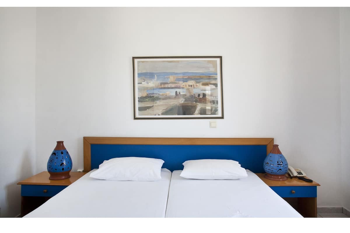 A double bed with white linens, a blue headboard, and two nightstands with decorative vases. A framed painting hangs above the bed.