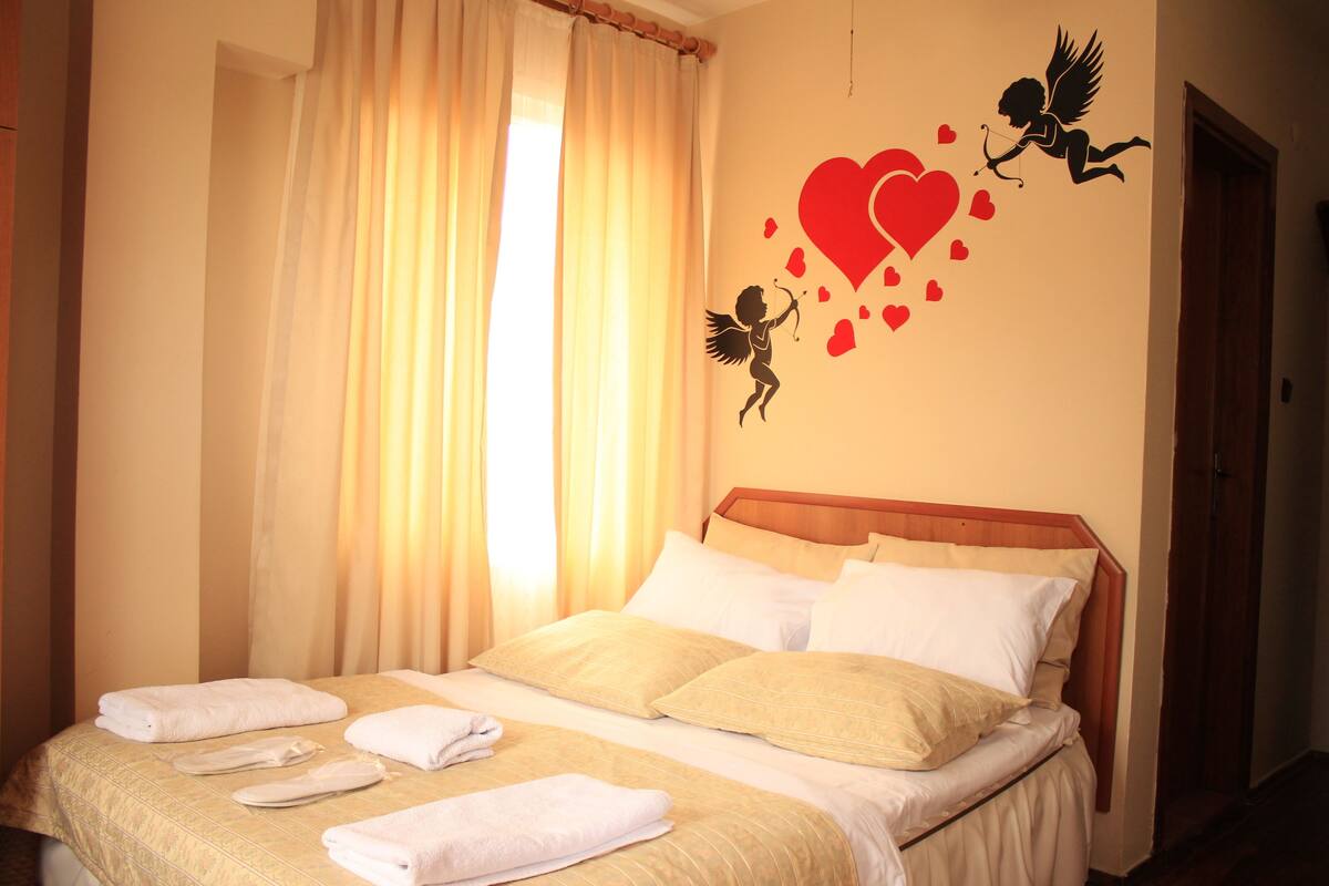 A hotel room with a bed, curtains, and a wall decoration of a cupid with hearts.