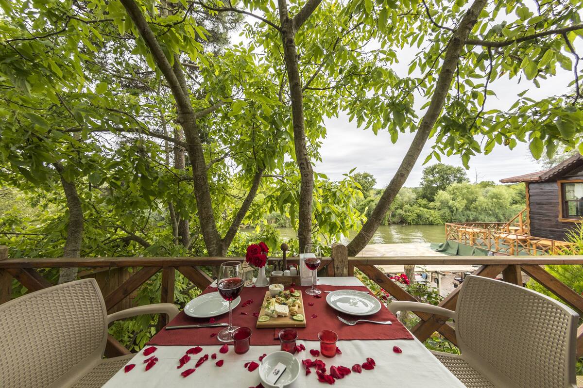 An outdoor dining area with a table set for two, surrounded by lush greenery and a view of a river.