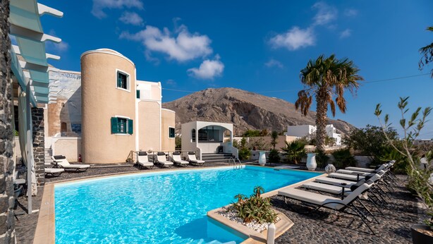 A swimming pool with lounge chairs, a mountain in the background, and a building with a curved facade. - Villa Anemomilos (Santorini)