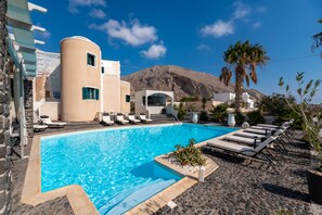 A swimming pool with lounge chairs, a mountain in the background, and a building with a curved facade. - Villa Anemomilos (Santorini)
