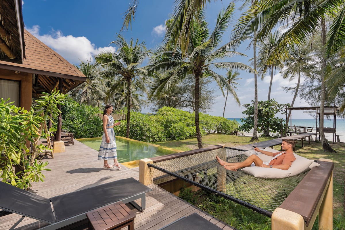 A wooden deck with a hammock, a pool, and palm trees.