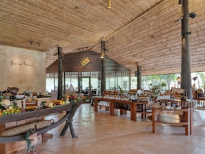 Enjoy meals in this open-plan restaurant with a high ceiling and large windows, featuring wooden tables and a central island with fresh produce.