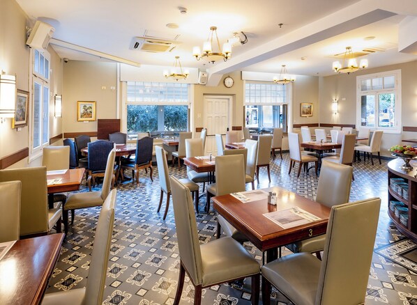 Un comedor de hotel con varias mesas y sillas, ventanas amplias e iluminación decorativa.