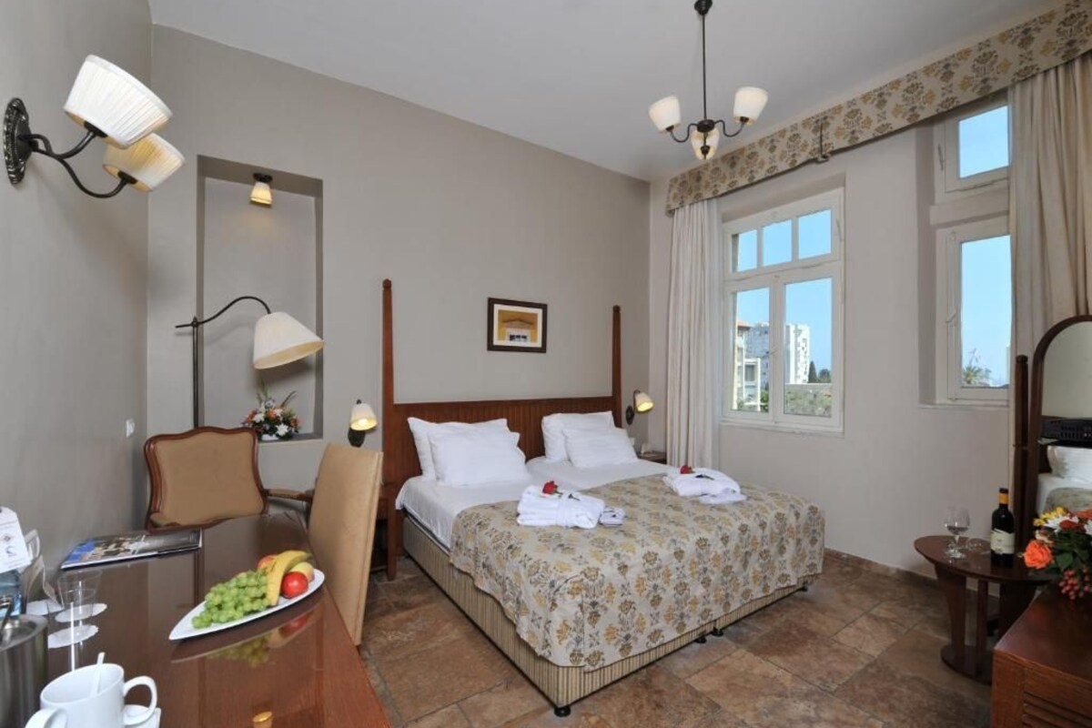 Habitación de hotel con cama, escritorio con silla, mesa de comedor y una fuente de frutas, y ventana con cortinas.