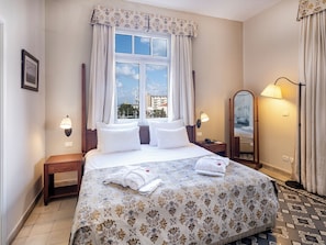 Habitación de hotel con una cama grande, dos mesitas de noche, un tocador, un espejo y una ventana con cortinas.