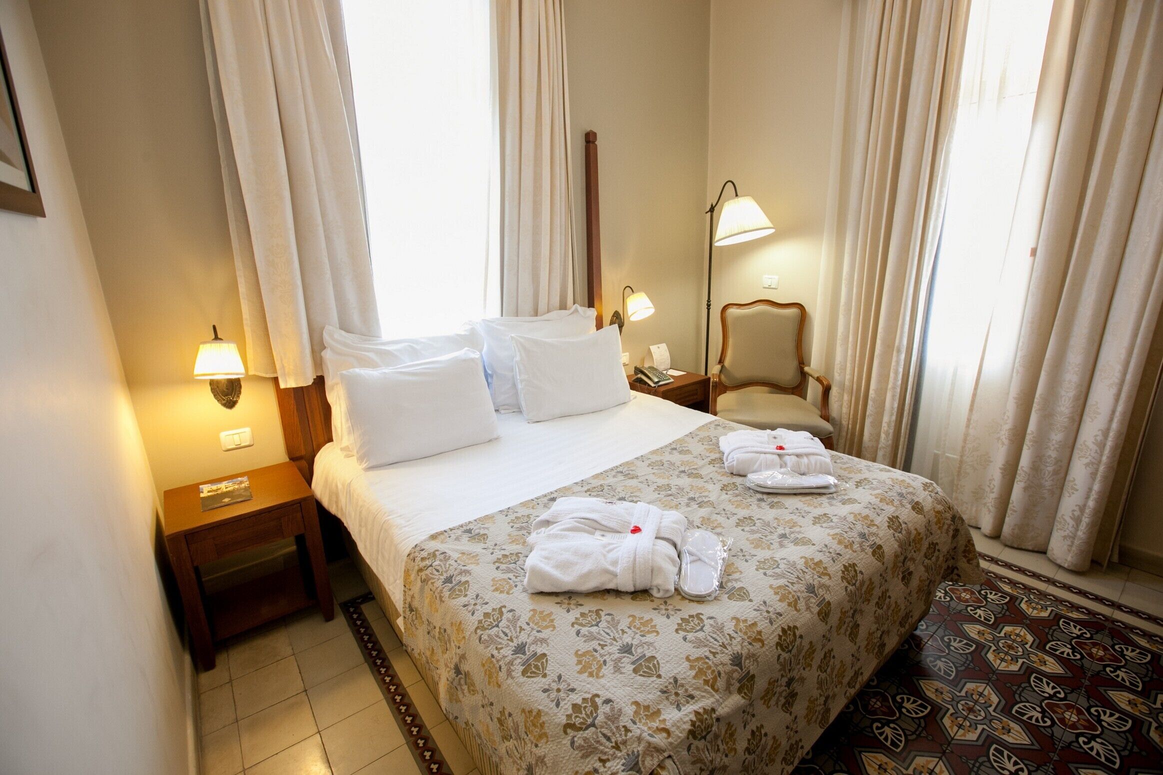 Un cuarto de hotel con una cama bien hecha, una mesita de noche con lámpara, una silla y un tapete estampado.