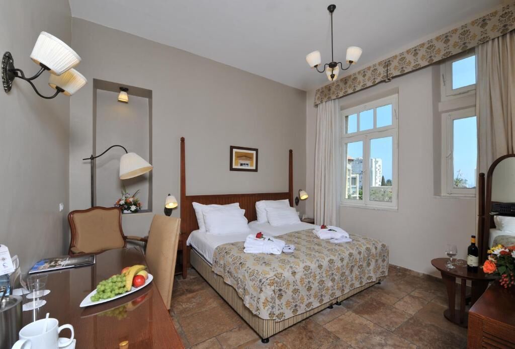 A hotel room with a bed, a desk with a chair, a dining table with a plate of fruit, and a window with curtains.