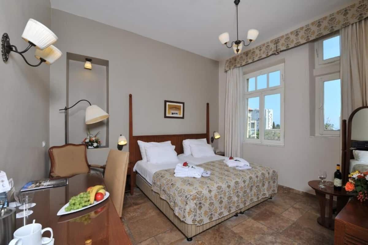 Habitación de hotel con cama, escritorio con silla, mesa de comedor y una fuente de frutas, y ventana con cortinas.