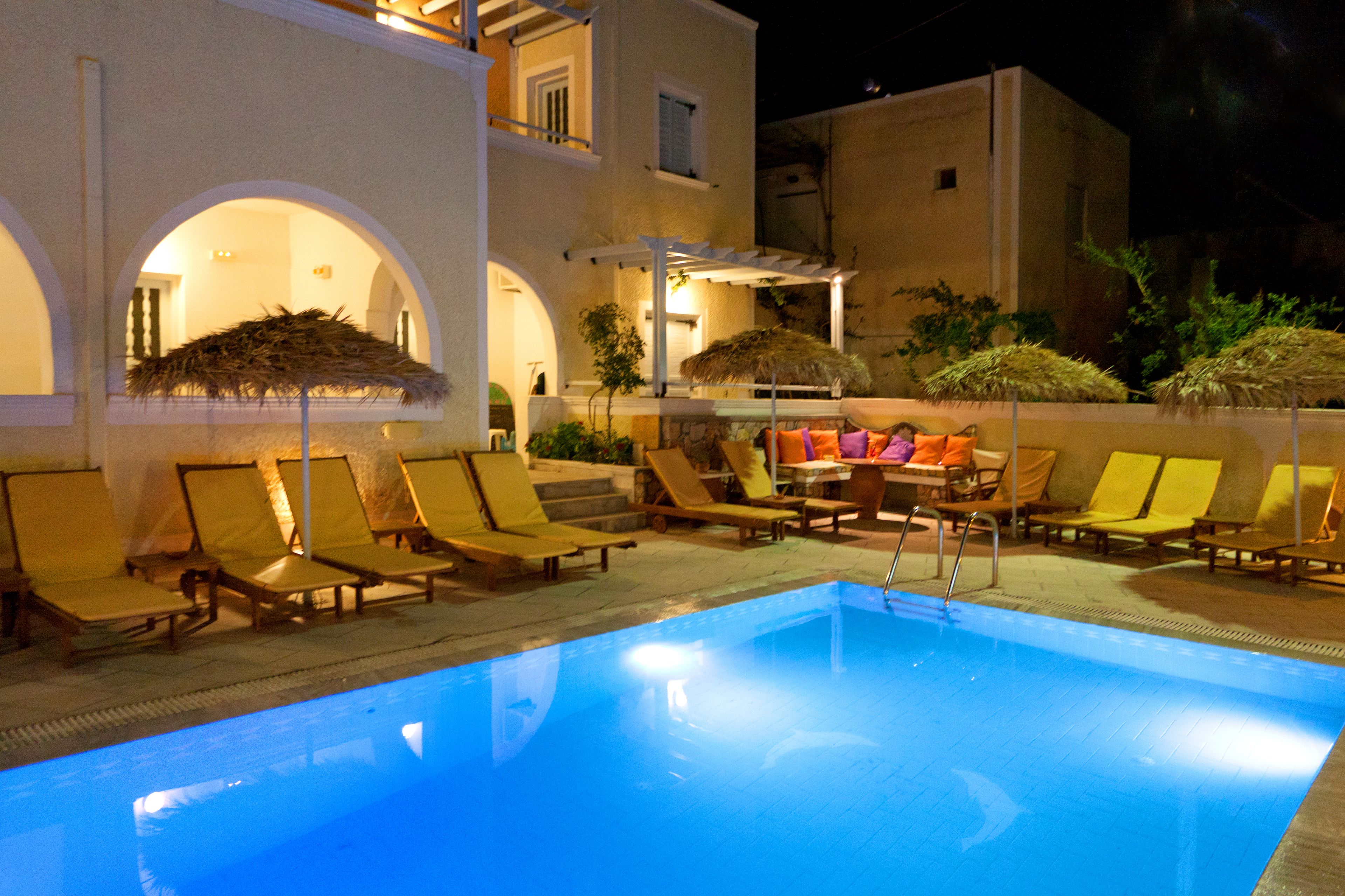 A swimming pool with lounge chairs and umbrellas, surrounded by buildings with arched windows and a thatched roof structure.