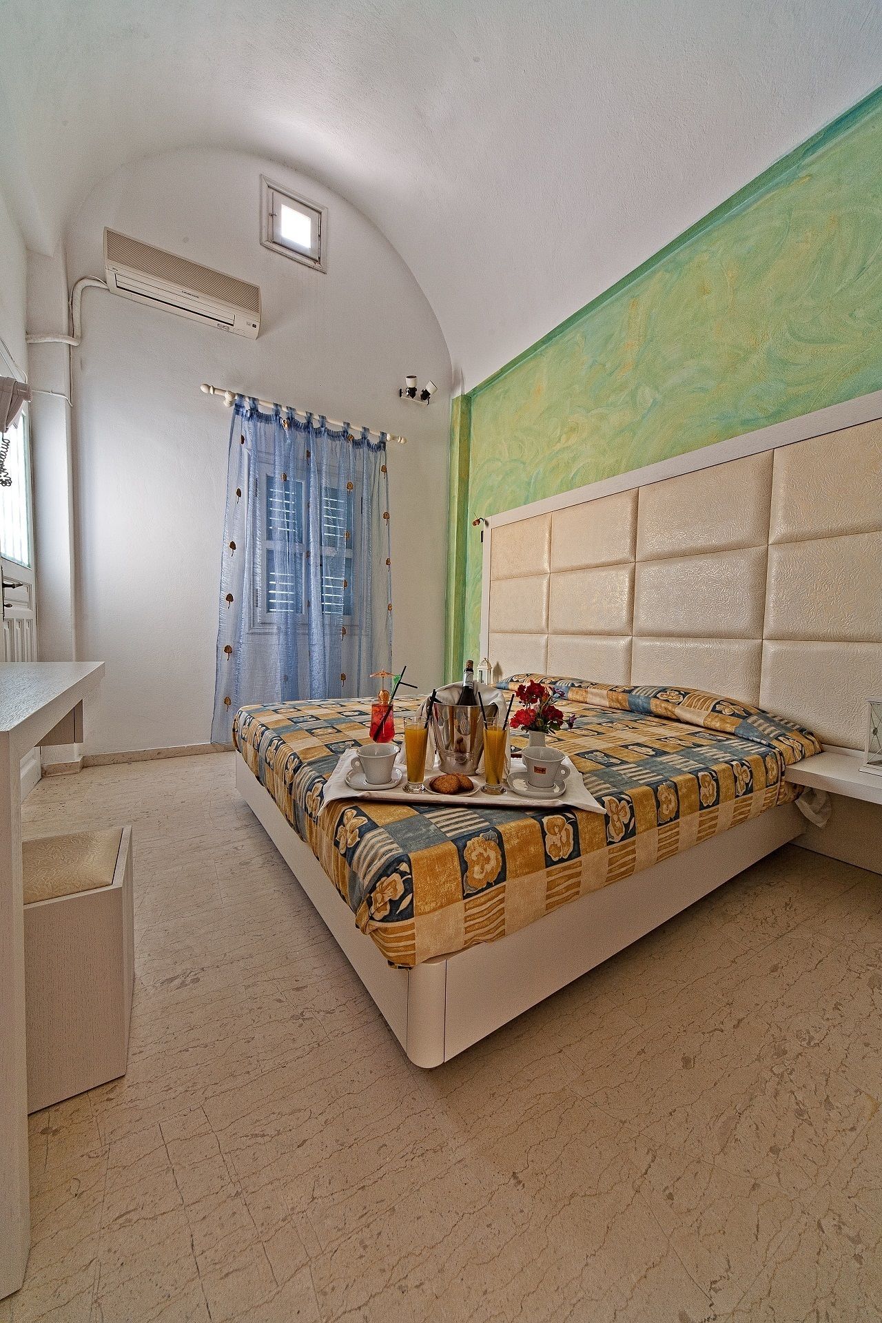 A bedroom with a bed, a bedside table, a desk, and a chair. There is a window with curtains, a wall-mounted air conditioner, and a green wall.