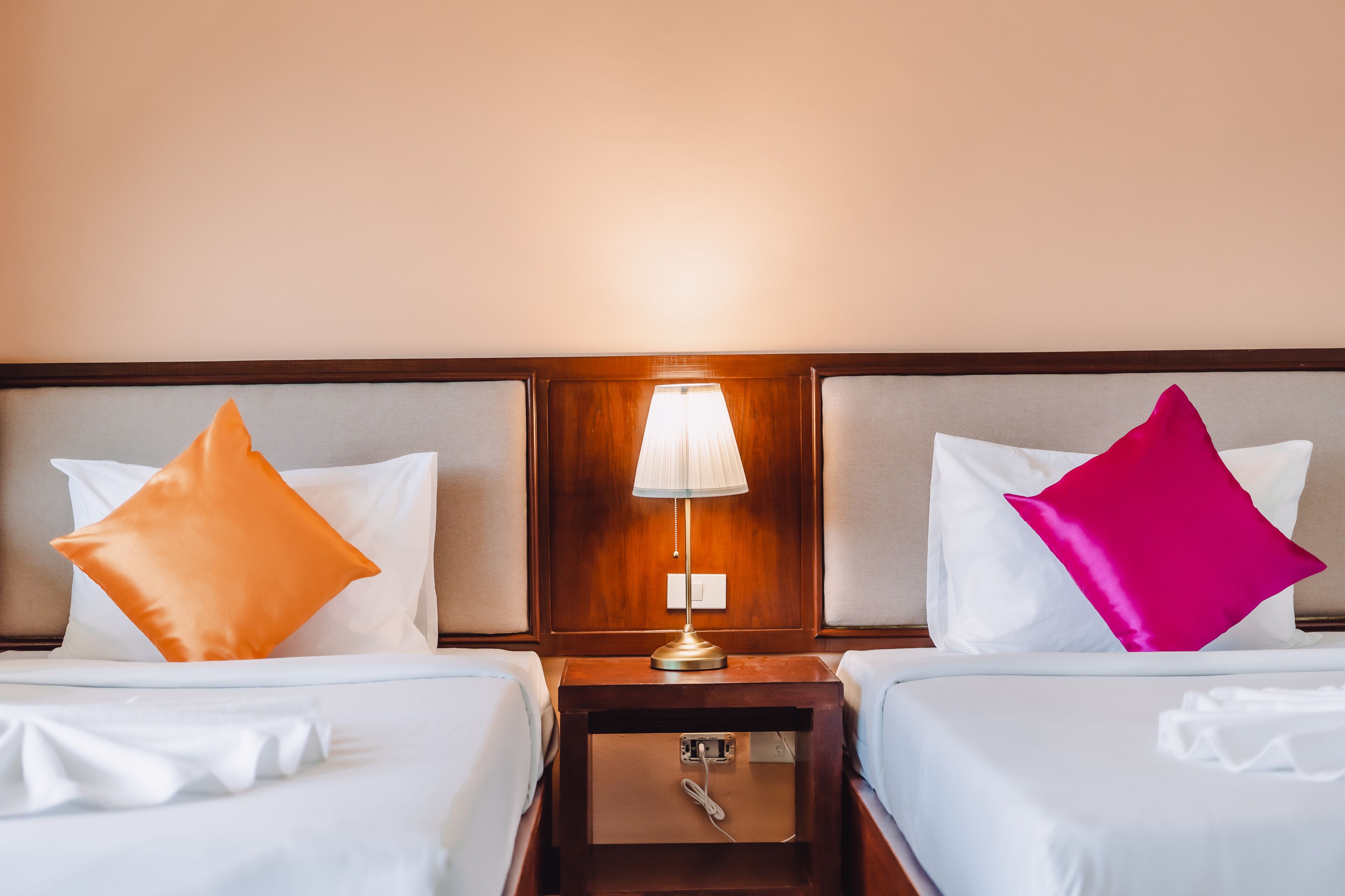 A hotel room with two beds, each with a white bedspread and a pillow. There is a wooden nightstand with a lamp between the beds.