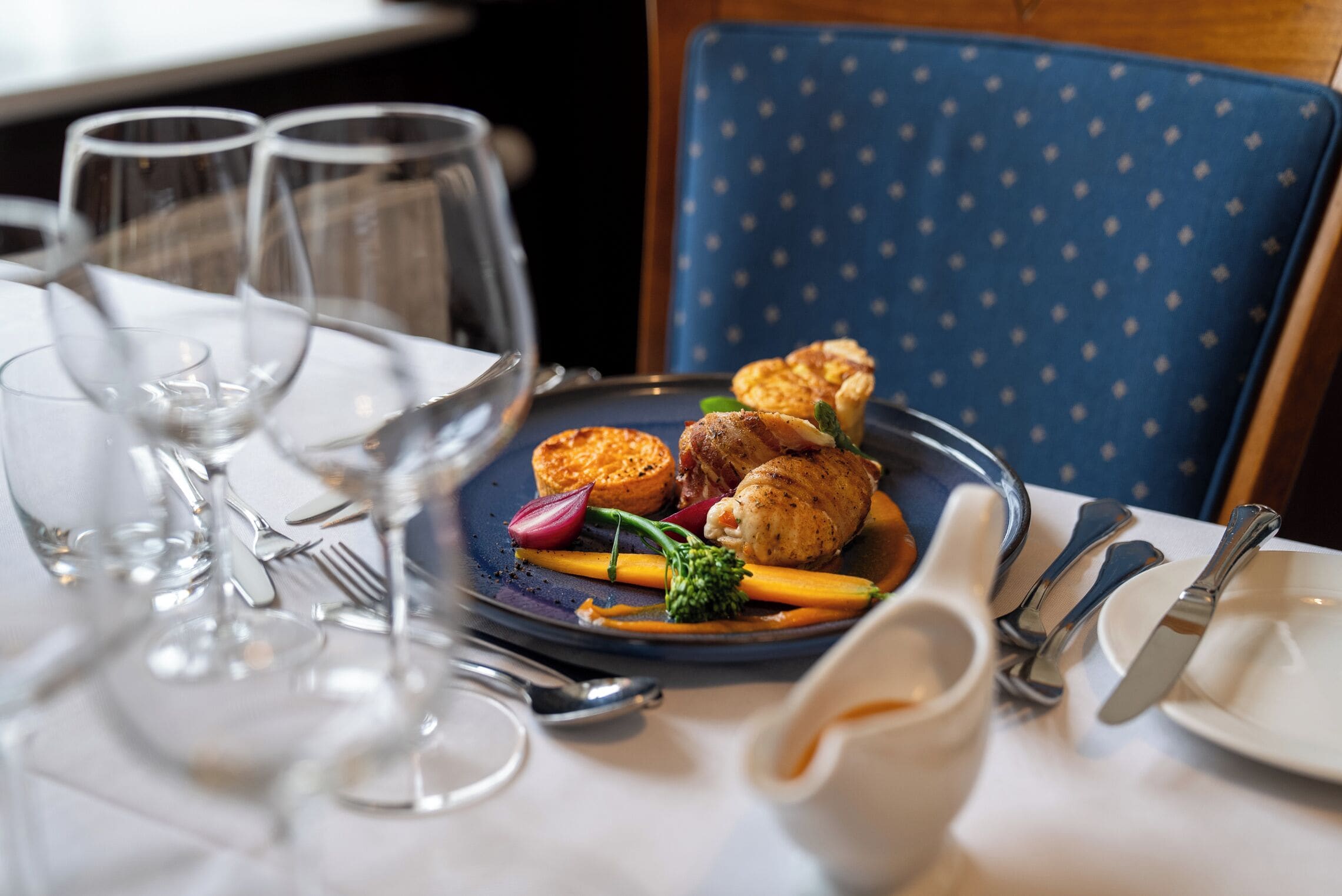 A dining table set with a plate of food, wine glasses, cutlery, and a blue chair.
