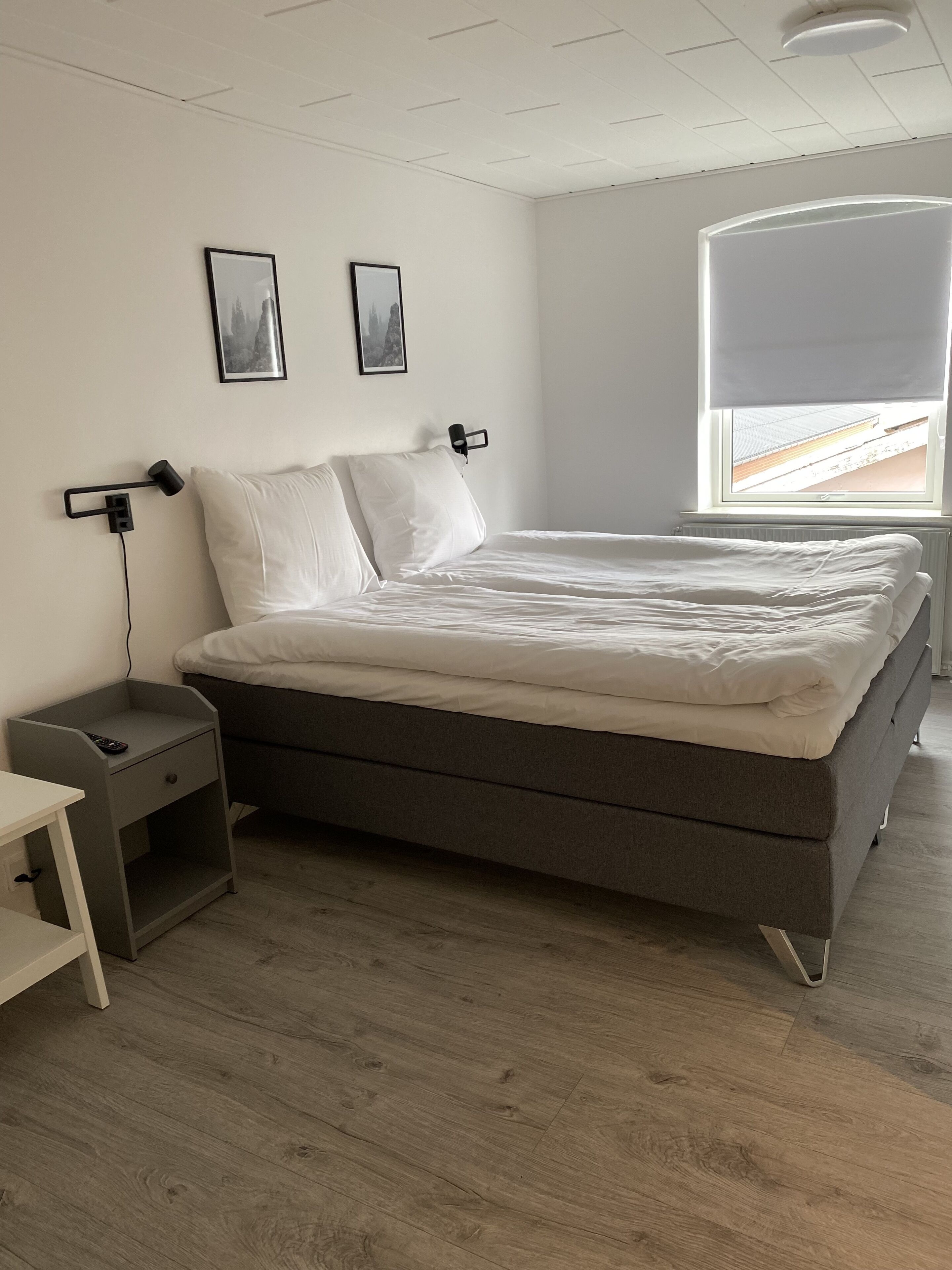 A modern bedroom with a grey bed, white bedding, a nightstand with a remote, and a window with a view of the outside.