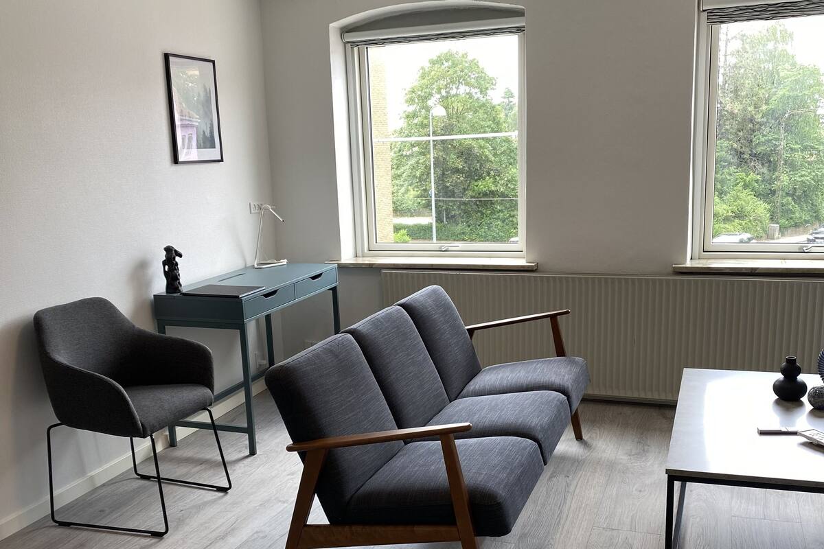 Una sala moderna con un sofá gris, una mesa de centro de madera y un escritorio con una silla. Hay dos ventanas que ofrecen luz natural y vistas a árboles.