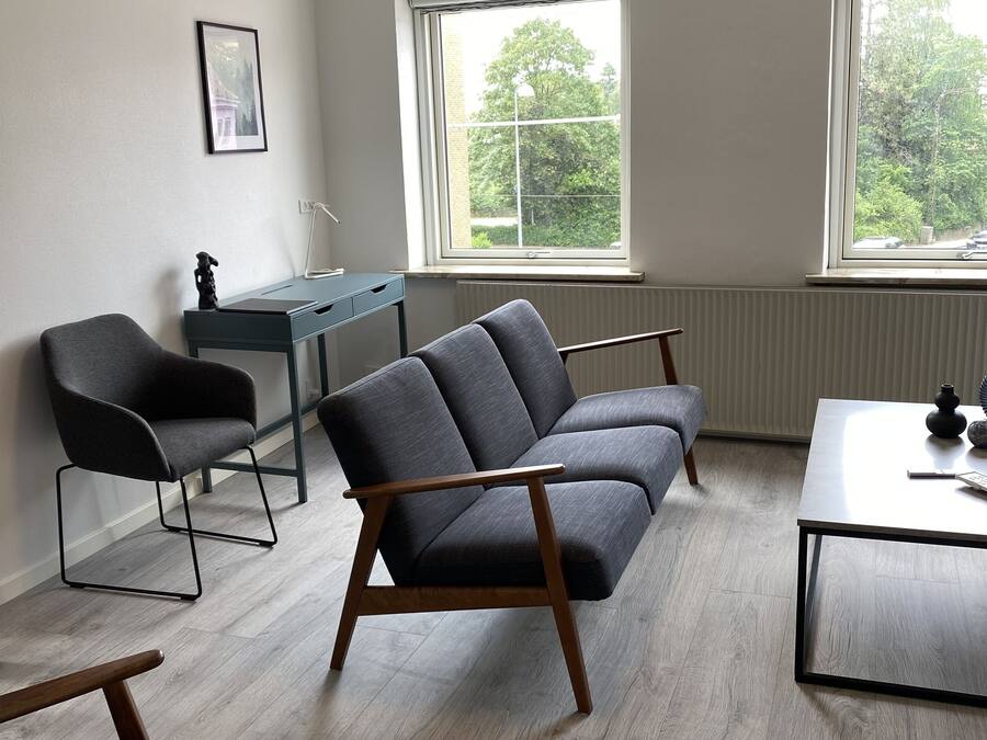 Un salon moderne avec un canapé gris, une table basse en bois et un bureau avec une chaise. Deux fenêtres laissent entrer la lumière naturelle et offrent une vue sur les arbres.