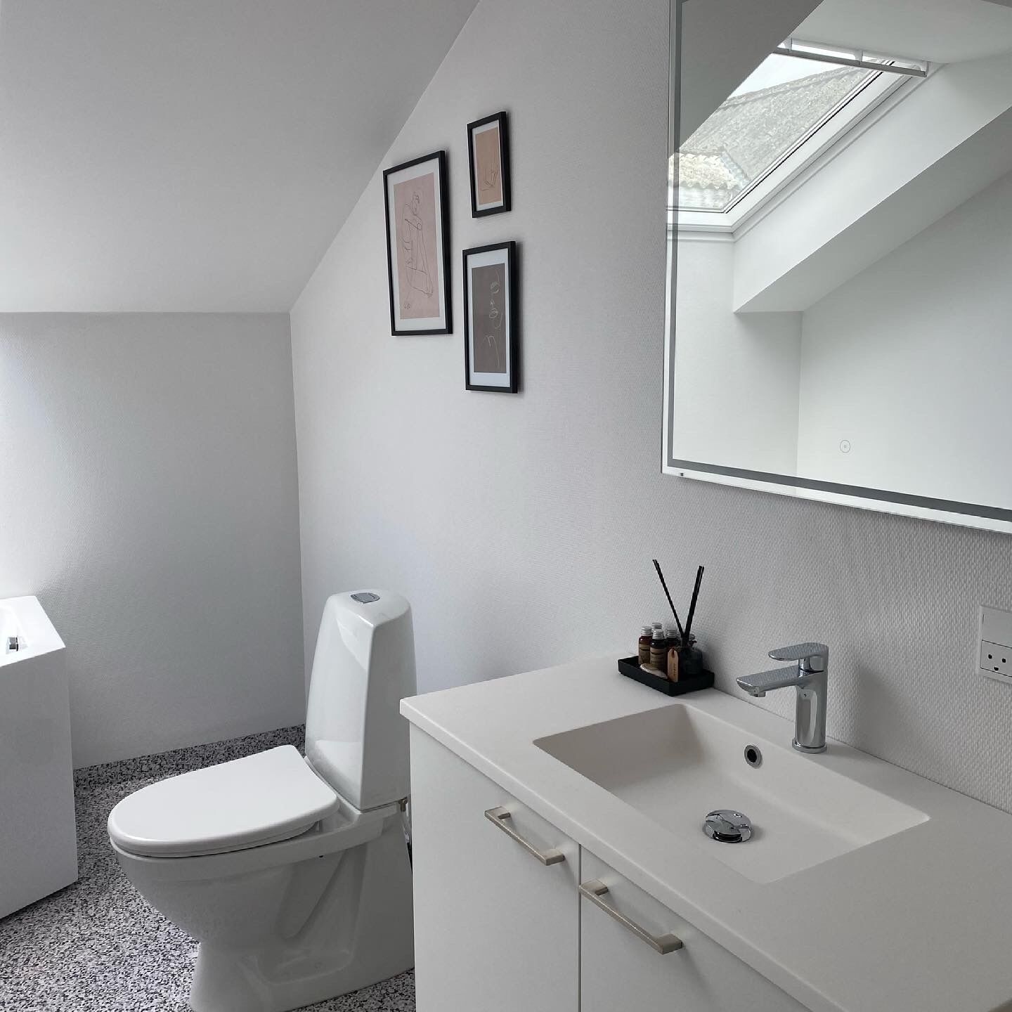 A modern bathroom with a toilet, sink, and mirror. There are framed pictures on the wall and a skylight above the mirror.
