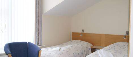 A hotel room with two beds, a wooden headboard, a blue armchair, and a window with blinds.