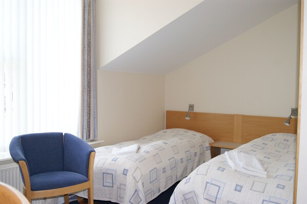 A hotel room with two beds, a wooden headboard, a blue armchair, and a window with blinds.