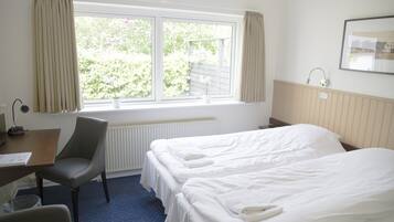 A hotel room with two beds, a desk, a chair, a window with curtains, a radiator, and a wall-mounted picture.