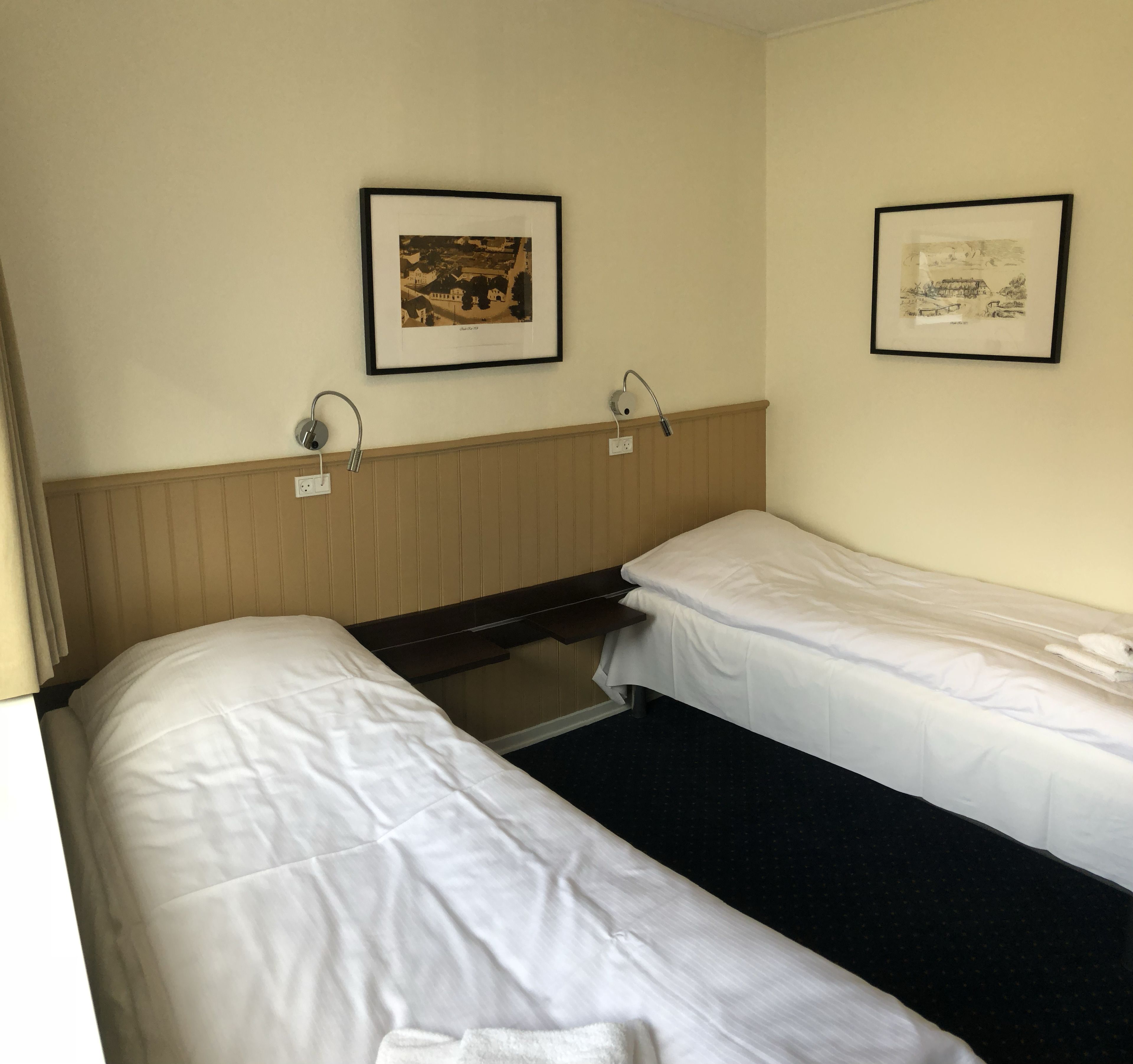 A hotel room with two single beds, a wooden headboard, a wall-mounted lamp, and two framed pictures.